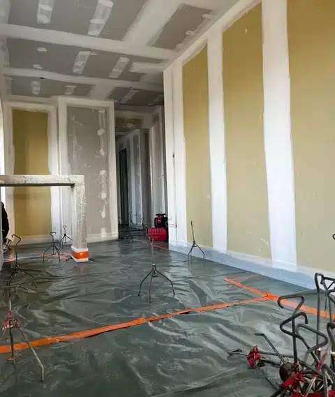 Intérieur en construction, murs en placo partiellement enduits, plafond brut. Sol couvert d'une bâche protectrice avec trépieds et ruban orange.