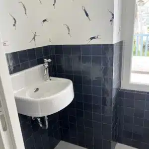 Petite salle d'eau avec lavabo mural blanc, carrelage métro noir brillant, et papier peint à motifs de nageurs plongeant.