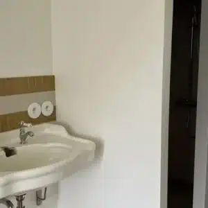 Salle de bain claire avec lavabo blanc vintage, robinetterie chromée et carreaux muraux moutarde/gris. Douche visible à droite.