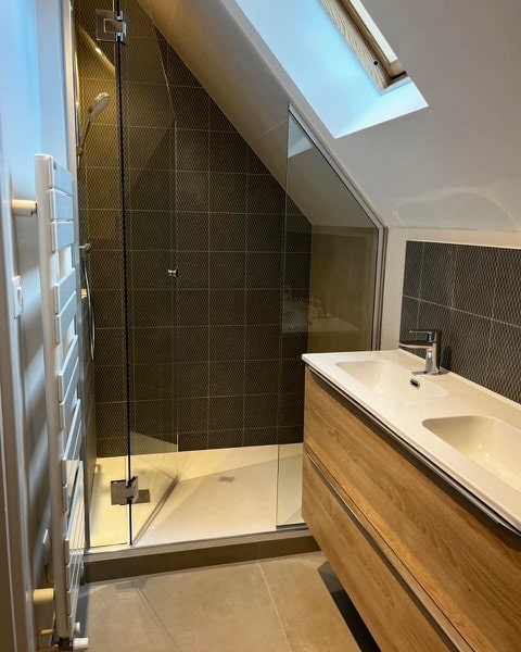 Salle de bain sous comble avec douche italienne, meuble bois et carrelage gris.